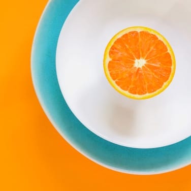Half of an orange in the center of a white plate, in the center of a light blue plate, placed on an orange background